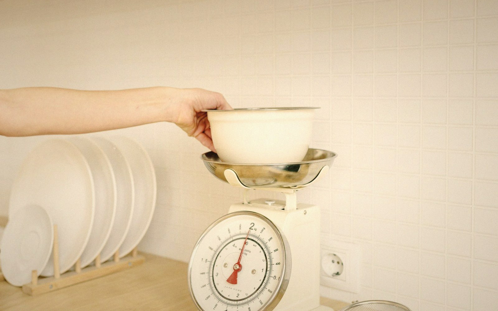 A hand places a bowl onto a kitchen scale beside a rack of stacked plates
